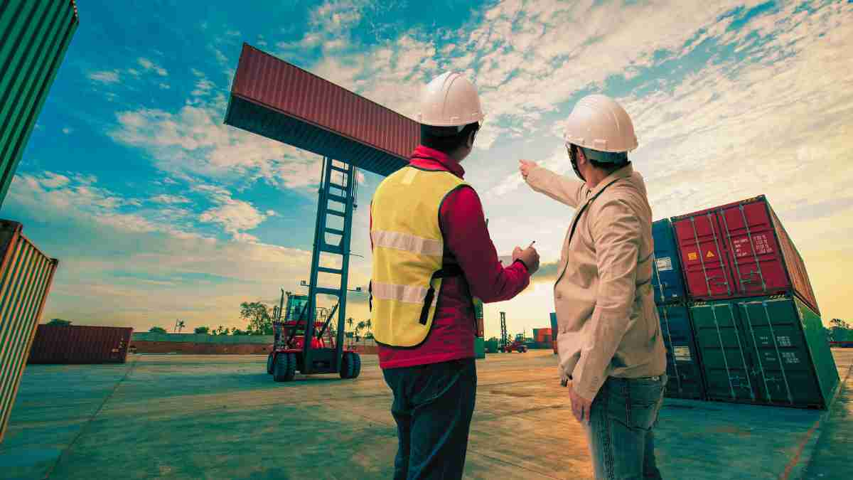 Two workers in safety gear overseeing the loading of shipping containers at a port, representing the concept of what is custom duty in international trade.